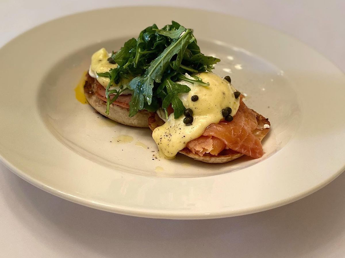 An elegant eggs Benedict rests on a white plate, layering a toasted English muffin with smoked salmon, poached eggs, and rich hollandaise dotted with capers. A bright tangle of dressed arugula tops the dish, with golden yolk pooling on the plate.