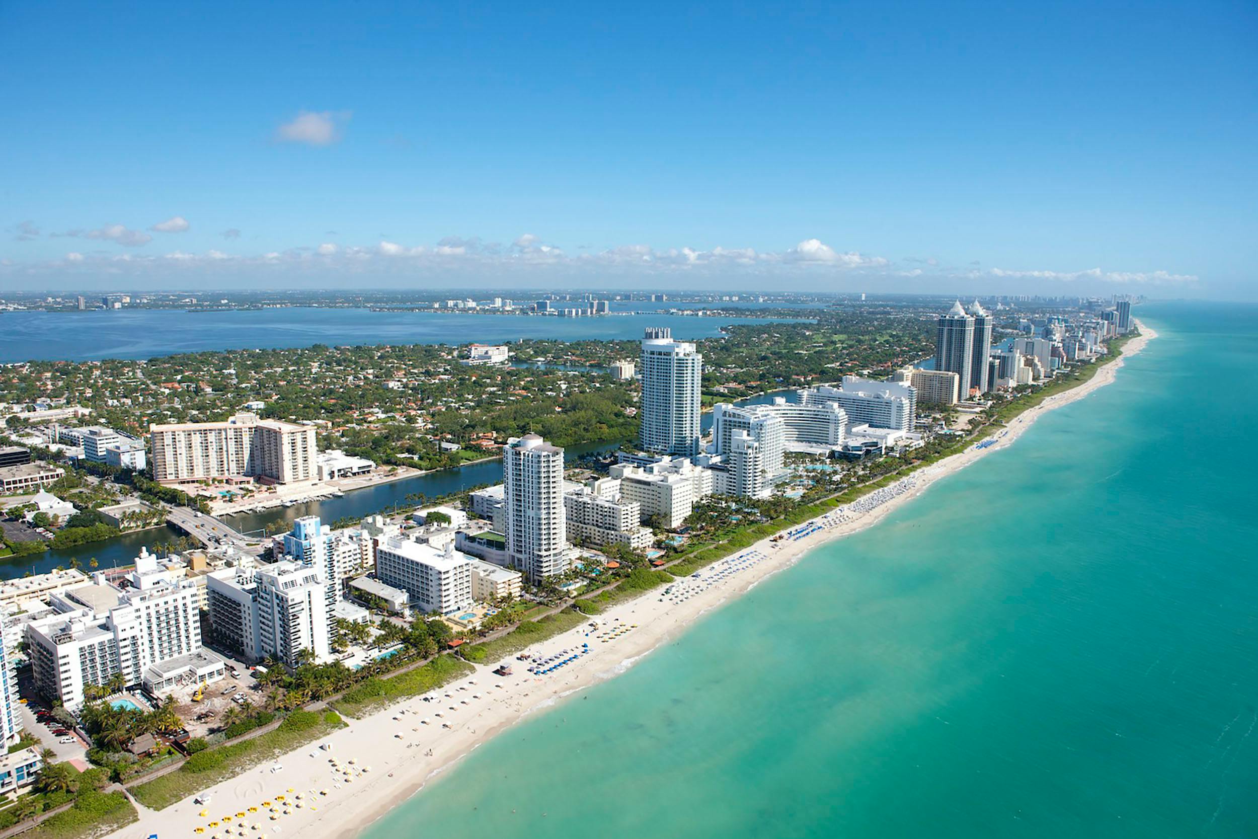 An aerial view shows Miami’s long sandy shoreline, bright turquoise ocean, and tall hotels lining the coast. The sky is clear and blue, giving the whole city a clean and sunny look. It captures the busy and beautiful energy of Miami Beach.