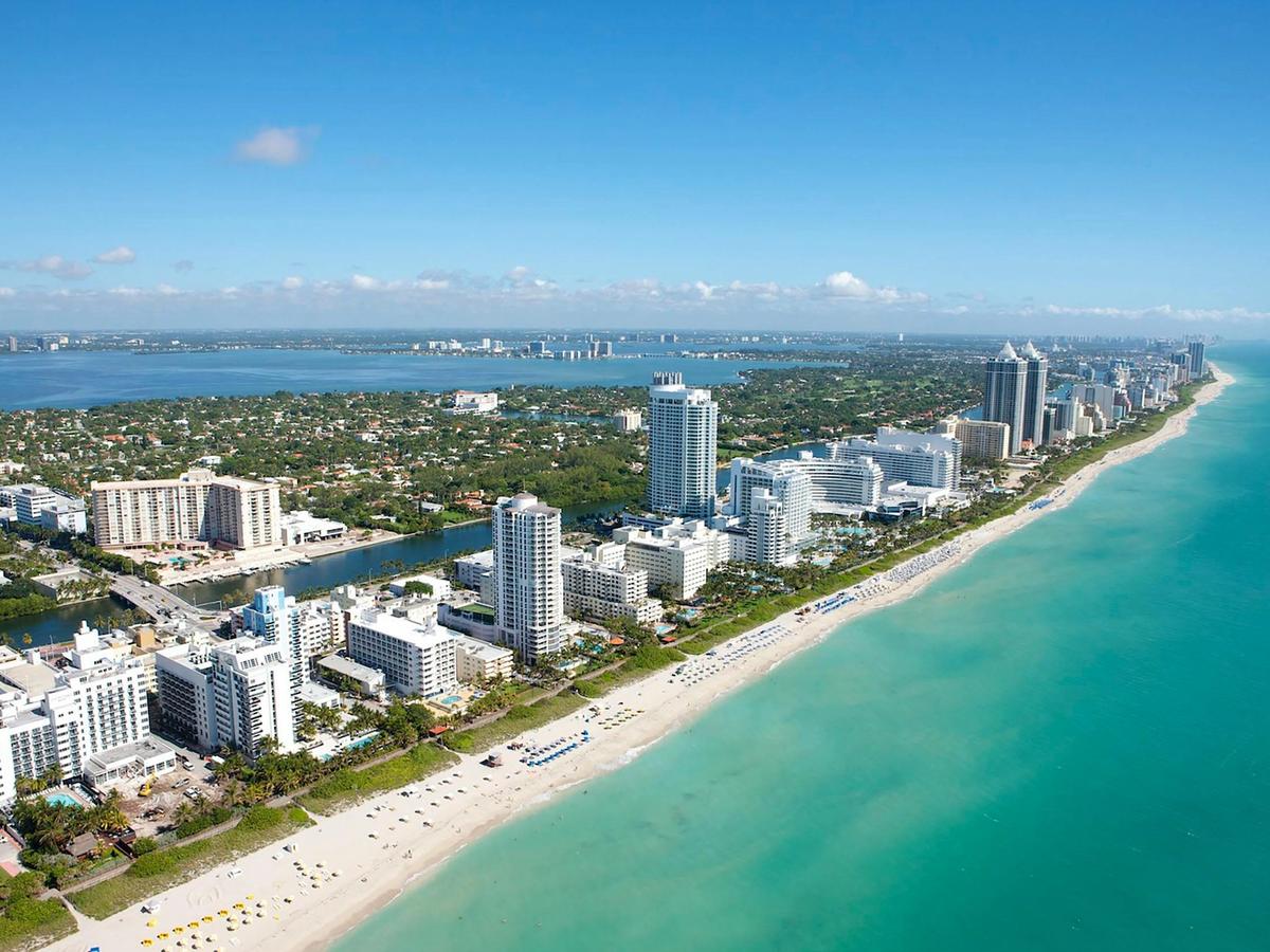 An aerial view shows Miami’s long sandy shoreline, bright turquoise ocean, and tall hotels lining the coast. The sky is clear and blue, giving the whole city a clean and sunny look. It captures the busy and beautiful energy of Miami Beach.