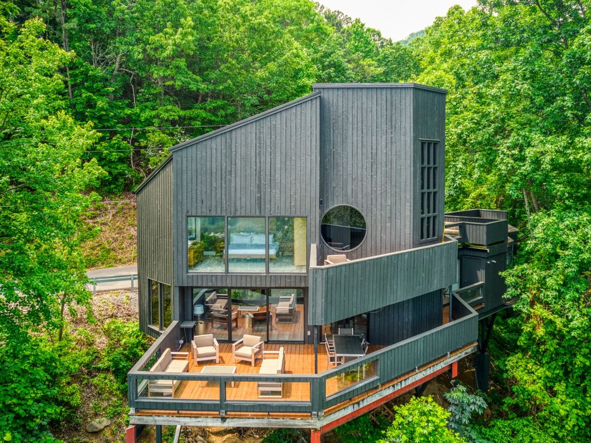 A modern mountain cabin built on stilts, surrounded by lush forest, with large windows and a spacious deck for outdoor lounging. Perfect for guests who want both luxury and an adventurous cliffside stay in the Smoky Mountains.
