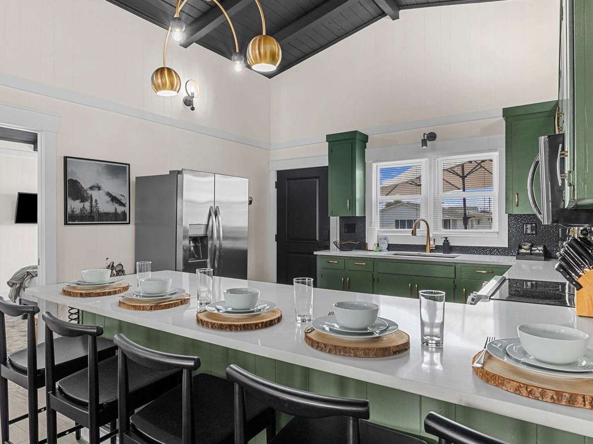 A modern kitchen with green cabinets, black accents, and a large island set with wood-slice chargers, white dishes, and tall glasses beneath gold pendant lights.