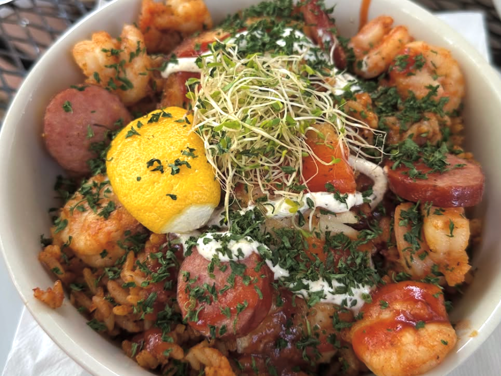 A bowl of jambalaya-style rice topped with shrimp, sausage slices, vegetables, fresh herbs, a lemon wedge, sprouts, and a drizzle of creamy sauce.