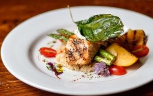 A beautifully plated gourmet dish featuring grilled seafood, roasted vegetables, and fresh herbs on a white plate. The colorful presentation highlights seasonal ingredients and careful preparation. This elegant dish reflects the refined dining experience at The Orchard restaurant.
