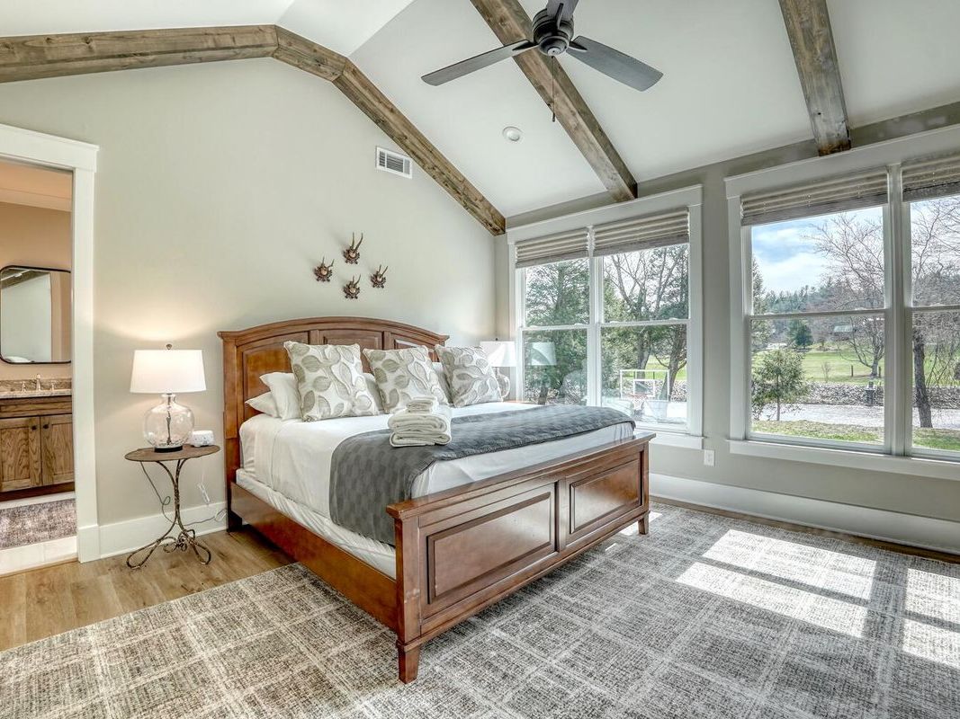 A bright and cozy bedroom features a wooden bed with plush pillows, vaulted ceilings with exposed beams, large windows with scenic views, and an attached bathroom.