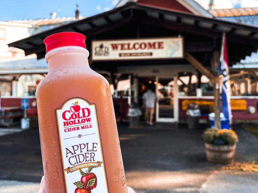 An apple cider drink is held up against the rustic backdrop of Cold Hollow Cider Mill. The sunshine and mountain views make this sweet Vermont treat taste even better after a day of exploring Stowe.