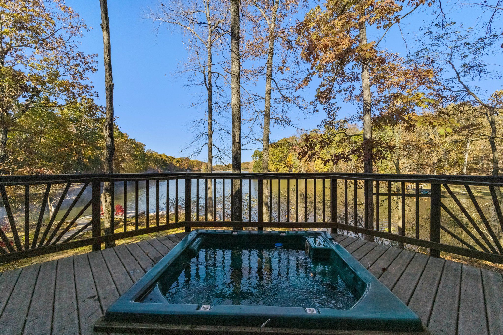 A built-in hot tub on a wooden deck overlooking a peaceful lake, surrounded by autumn trees and framed by a decorative wooden railing under a clear blue sky.