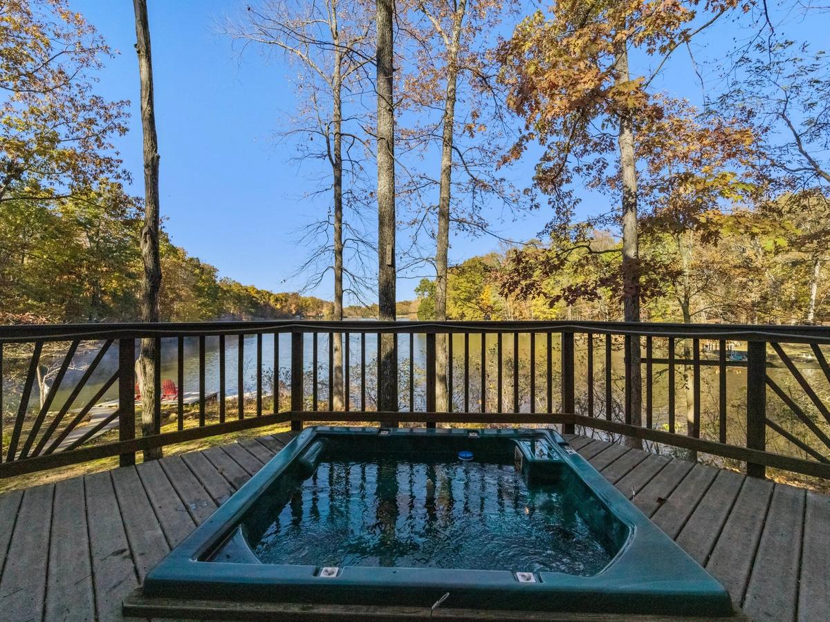 A built-in hot tub on a wooden deck overlooking a peaceful lake, surrounded by autumn trees and framed by a decorative wooden railing under a clear blue sky.