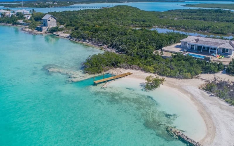 A private beachfront estate in the Bahamas with clear turquoise water, white sand, and a wooden dock leading into the sea. The luxury villa sits along a quiet shoreline surrounded by lush greenery and calm island views. Sueno De Mar offers a peaceful oceanfront escape perfect for a tropical getaway.