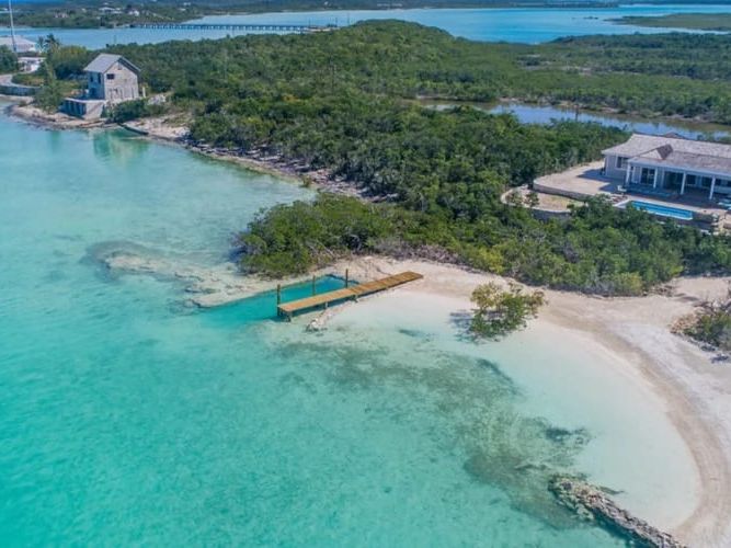 A private beachfront estate in the Bahamas with clear turquoise water, white sand, and a wooden dock leading into the sea. The luxury villa sits along a quiet shoreline surrounded by lush greenery and calm island views. Sueno De Mar offers a peaceful oceanfront escape perfect for a tropical getaway.