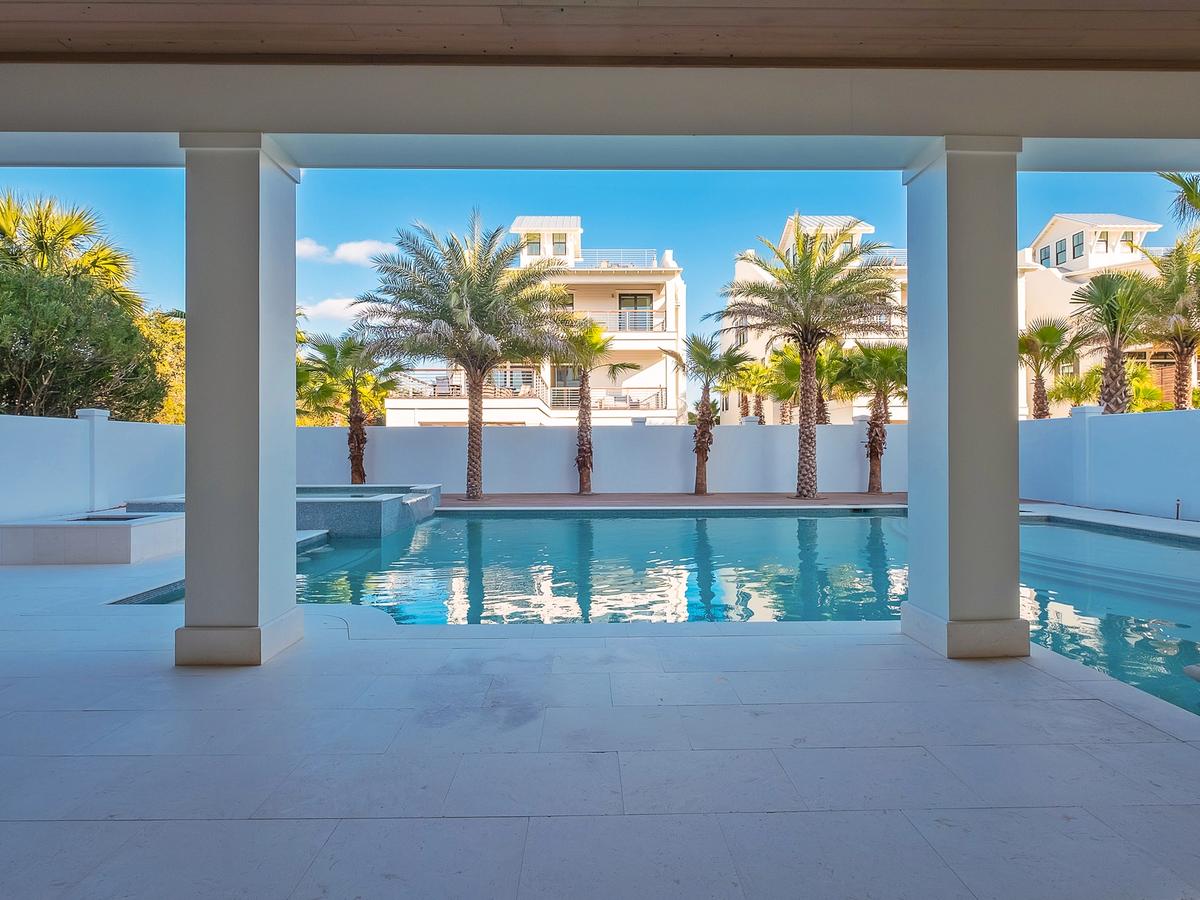 A covered patio overlooks a serene swimming pool surrounded by palm trees and modern white buildings under a bright blue sky.