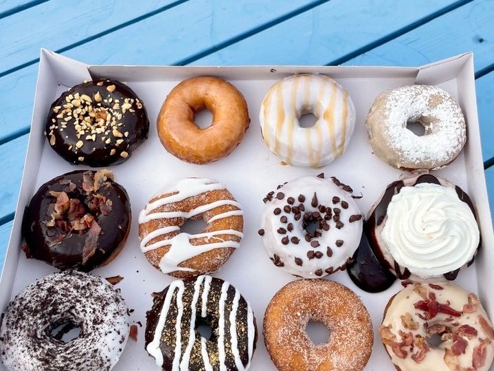 A colorful box of 12 custom donuts from Duck Donuts, each topped with different goodies like bacon, sprinkles, chocolate drizzle, and powdered sugar. This hand-held treat box is ready to be devoured on a sunny day at a blue picnic table.