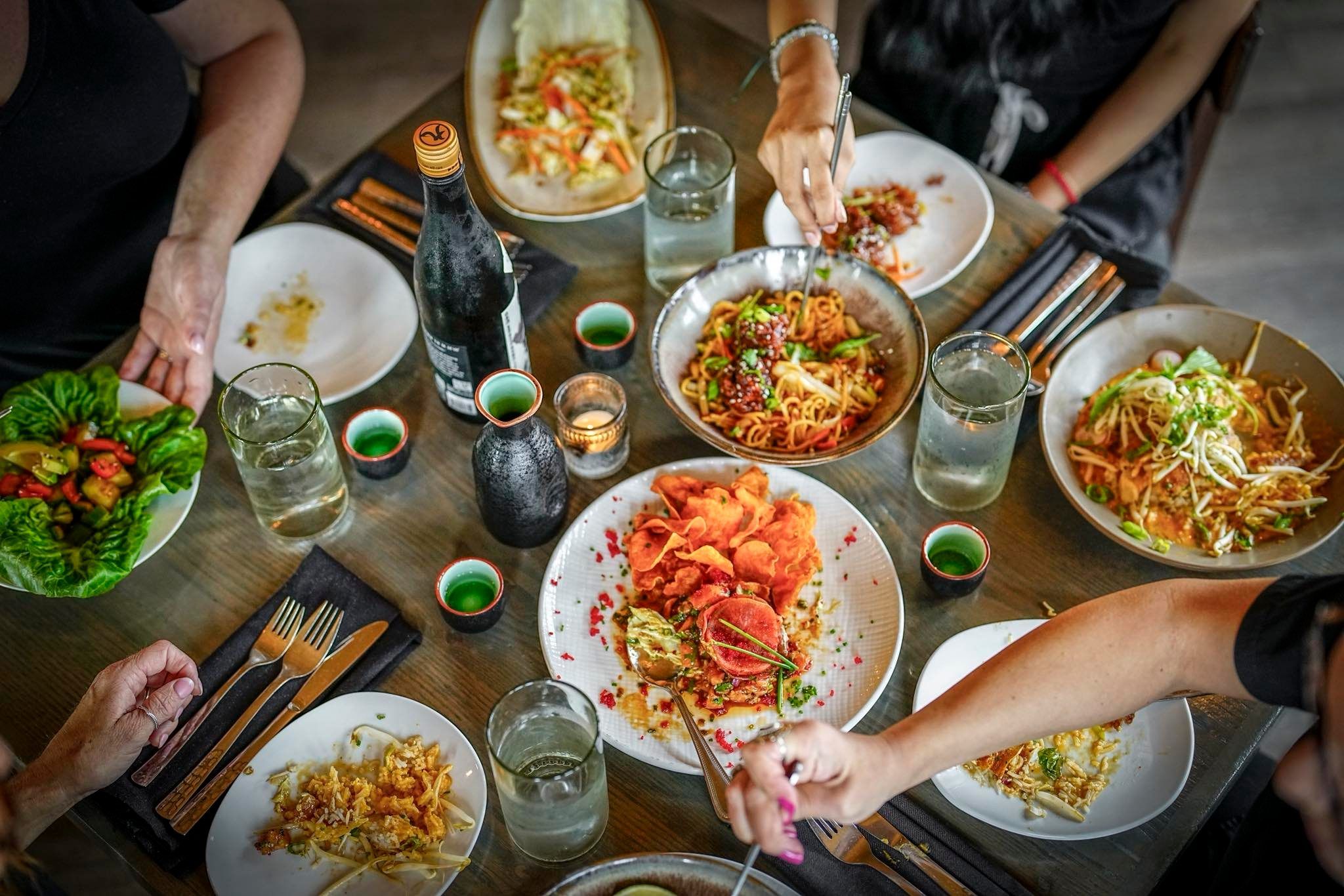 Friends gather around a table sharing colorful noodle dishes, appetizers, and drinks. The lively scene captures the social experience of enjoying Asian-inspired cuisine together. The Stubborn Buddha offers a vibrant dining experience with creative flavors.