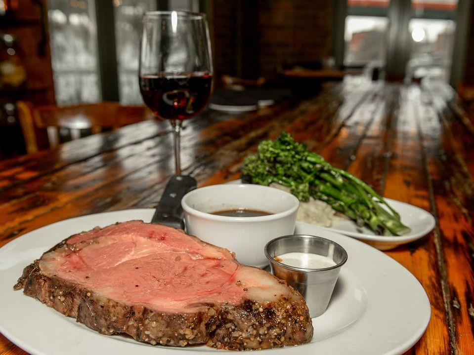 A thick cut of prime rib paired with broccolini, mashed potatoes, and red wine on a rustic wood table. Perfect steakhouse meal for meat lovers.