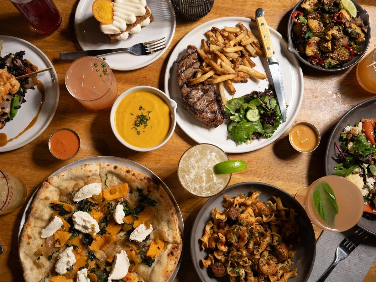 A table full of dishes at True Food Kitchen, including pizza, steak with fries, pasta, salads, and cocktails. The menu offers a balance of indulgent and wholesome options in a lively dining settin