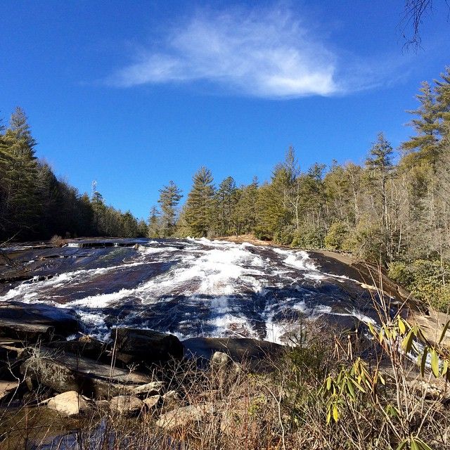 Water flows smoothly over the wide rock face at Bridal Veil Falls, surrounded by tall trees and clear blue skies. This scenic waterfall in North Carolina is a popular stop for visitors looking to enjoy easy access views and peaceful mountain scenery.