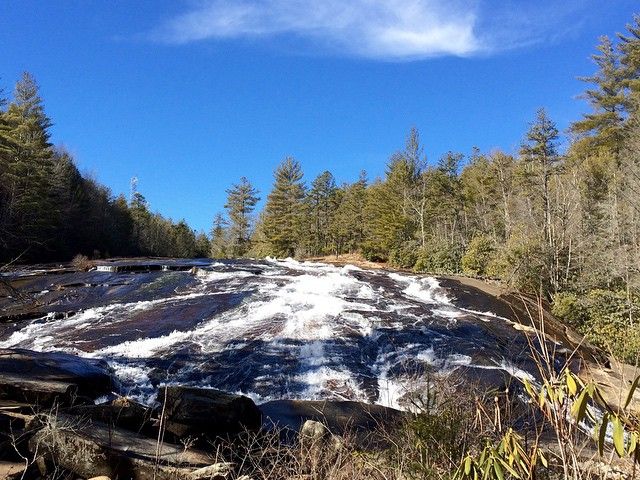Water flows smoothly over the wide rock face at Bridal Veil Falls, surrounded by tall trees and clear blue skies. This scenic waterfall in North Carolina is a popular stop for visitors looking to enjoy easy access views and peaceful mountain scenery.