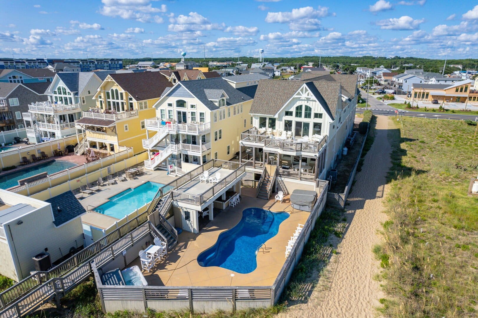 "A large, luxurious beachfront home with a private fenced-in pool area, surrounded by neighboring vacation rentals. The house features multiple levels of decks with outdoor seating, large windows, and a coastal-inspired design with a mix of soft yellow and cream exteriors. The backyard includes a curvy swimming pool, a hot tub, and multiple lounge areas with tables and chairs. A sandy path leads from the property to the beach, while the surrounding area is filled with other high-end beach homes and resort-style pools under a bright blue sky with scattered clouds.