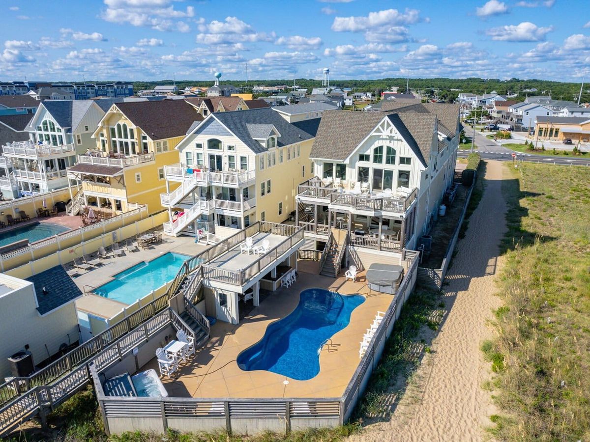 "A large, luxurious beachfront home with a private fenced-in pool area, surrounded by neighboring vacation rentals. The house features multiple levels of decks with outdoor seating, large windows, and a coastal-inspired design with a mix of soft yellow and cream exteriors. The backyard includes a curvy swimming pool, a hot tub, and multiple lounge areas with tables and chairs. A sandy path leads from the property to the beach, while the surrounding area is filled with other high-end beach homes and resort-style pools under a bright blue sky with scattered clouds.