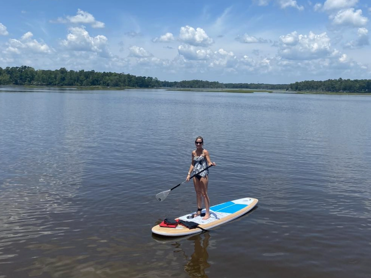 A woman in a swimsuit standing on a paddleboard with a paddle in hand, enjoying a sunny day on a calm lake surrounded by trees and a blue sky with scattered clouds.