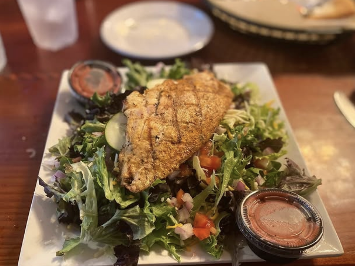 Grilled seasoned fish fillet served atop a fresh garden salad with mixed greens, diced tomatoes, cucumbers, and a side of vinaigrette dressing.