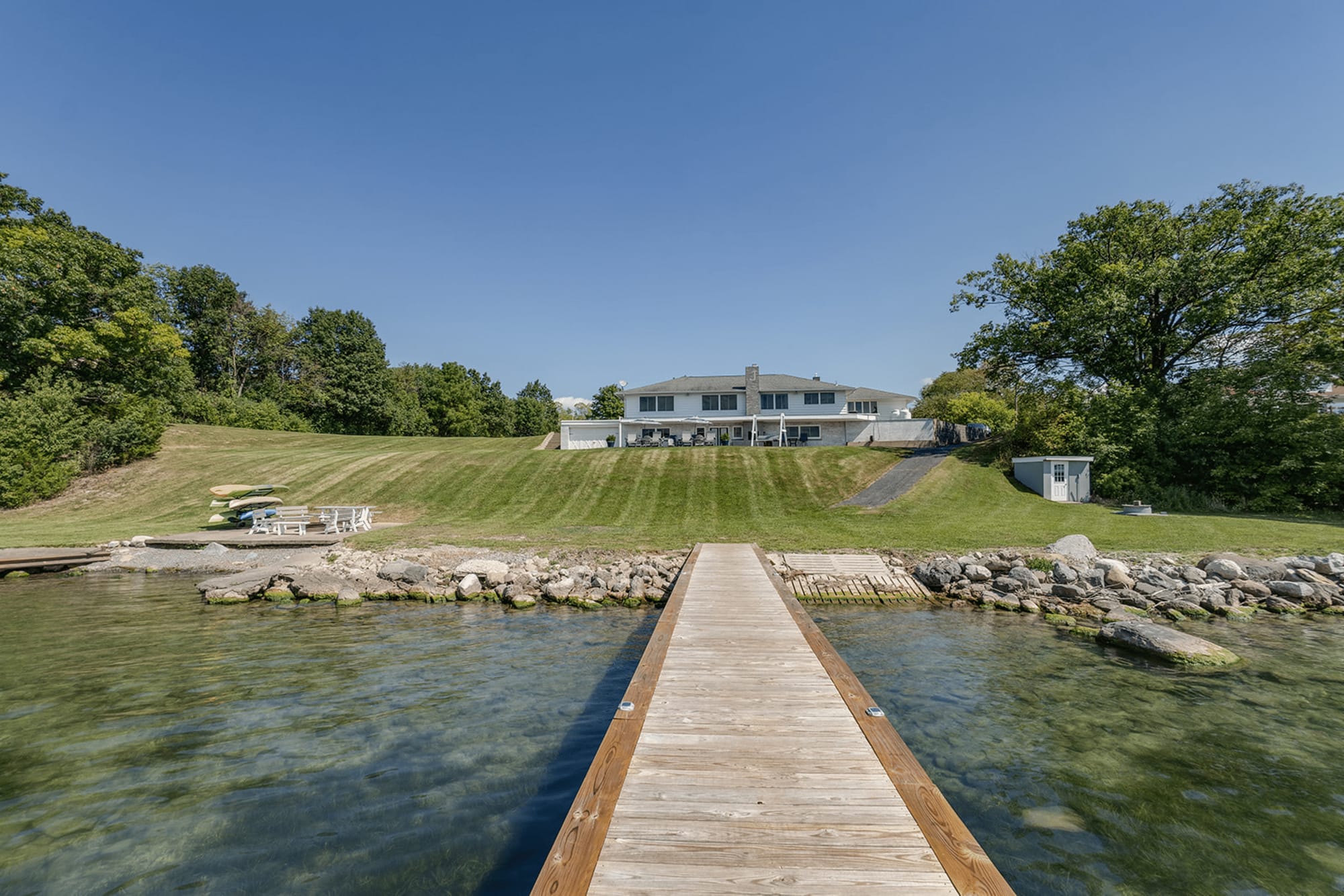 A tranquil Finger Lakes retreat with a private dock stretching over clear blue water. The open lawn and sunny skies make this home ideal for lakeside mornings and family gatherings.