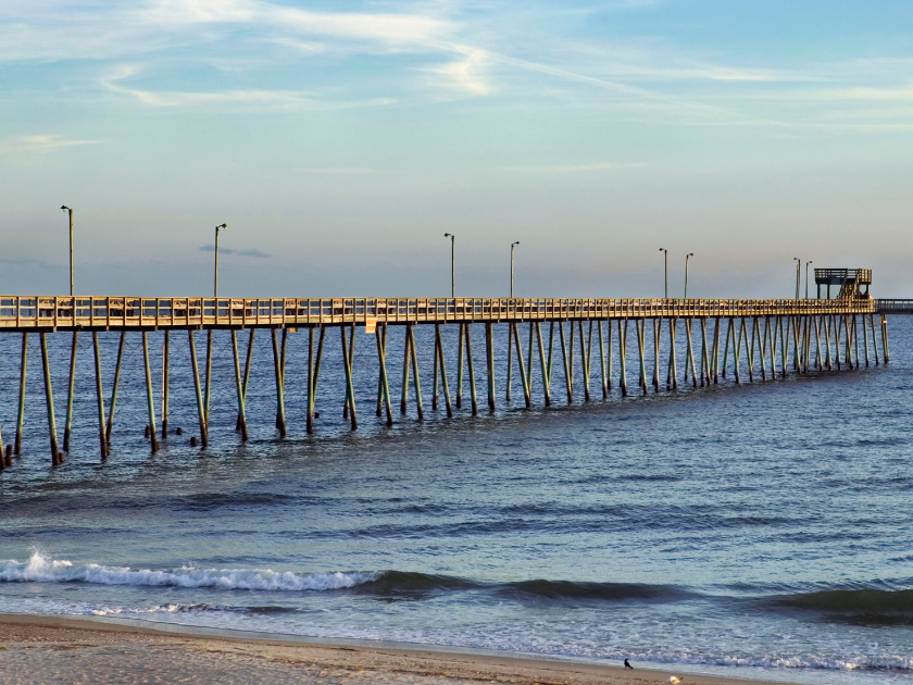 The morning sky glows gold behind a long wooden pier stretching out into the calm ocean. A peaceful way to start the day in Holden Beach.
