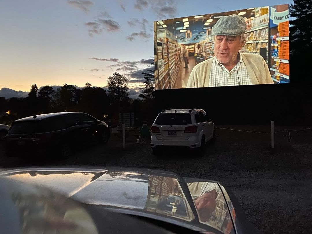 Cars are parked under the evening sky at a classic drive-in theater, with a movie playing on a giant outdoor screen. The warm glow of sunset and the nostalgic setting make it a cozy night out. Families relax in their vehicles while enjoying the film.