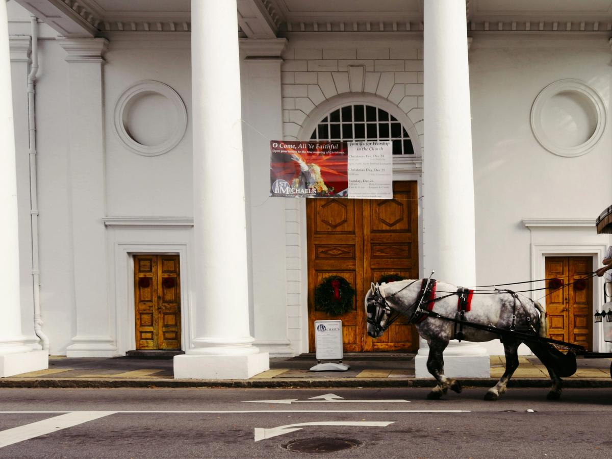 A horse-drawn carriage passes in front of the historic white-columned St. Michael’s Church in Charleston, South Carolina, with people seated inside enjoying a tour.