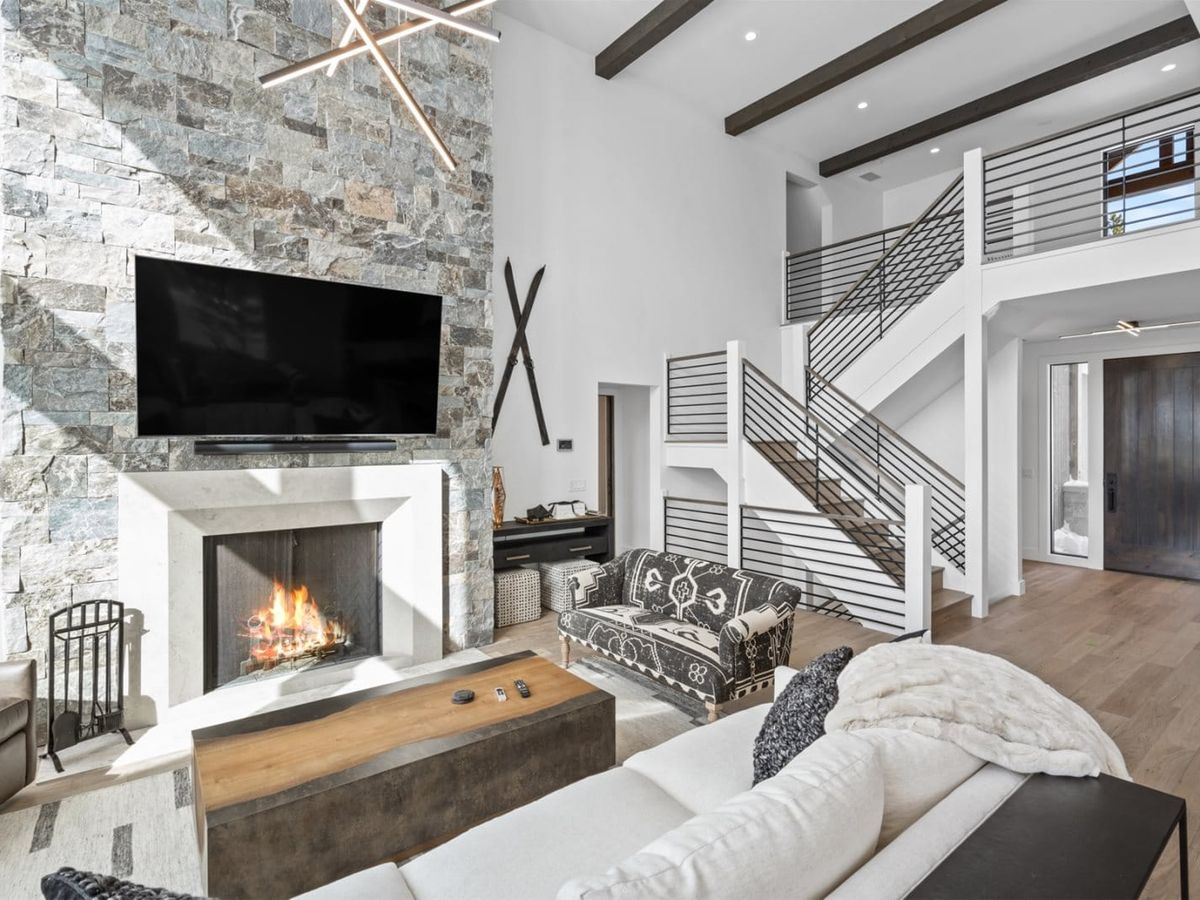A steaming hot tub sits on a covered wooden deck beside a stone cabin wall, with snowy trees and rustic wooden chairs in the background.