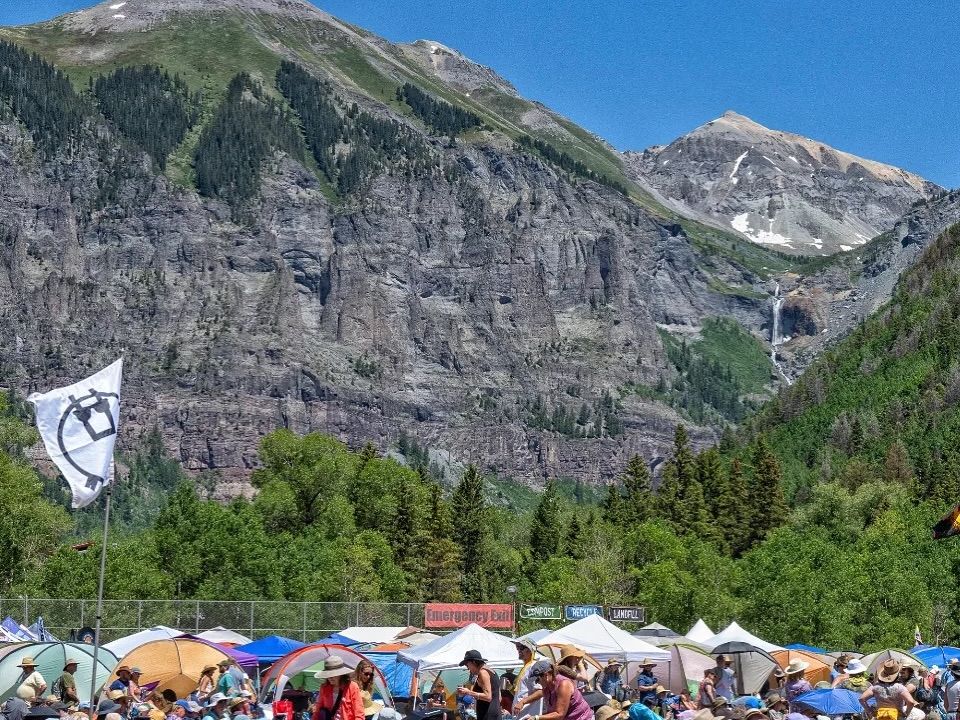 Crowds gather on a sunny day in Telluride, surrounded by tents and mountains, for the popular Bluegrass Festival. Music, nature, and community meet in this iconic Colorado event.