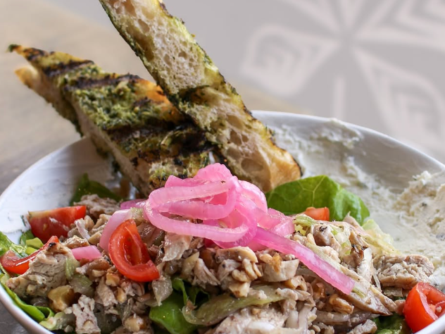 A fresh salad topped with shredded chicken, cherry tomatoes, walnuts, and pickled onions, served with pesto-grilled bread on the side.