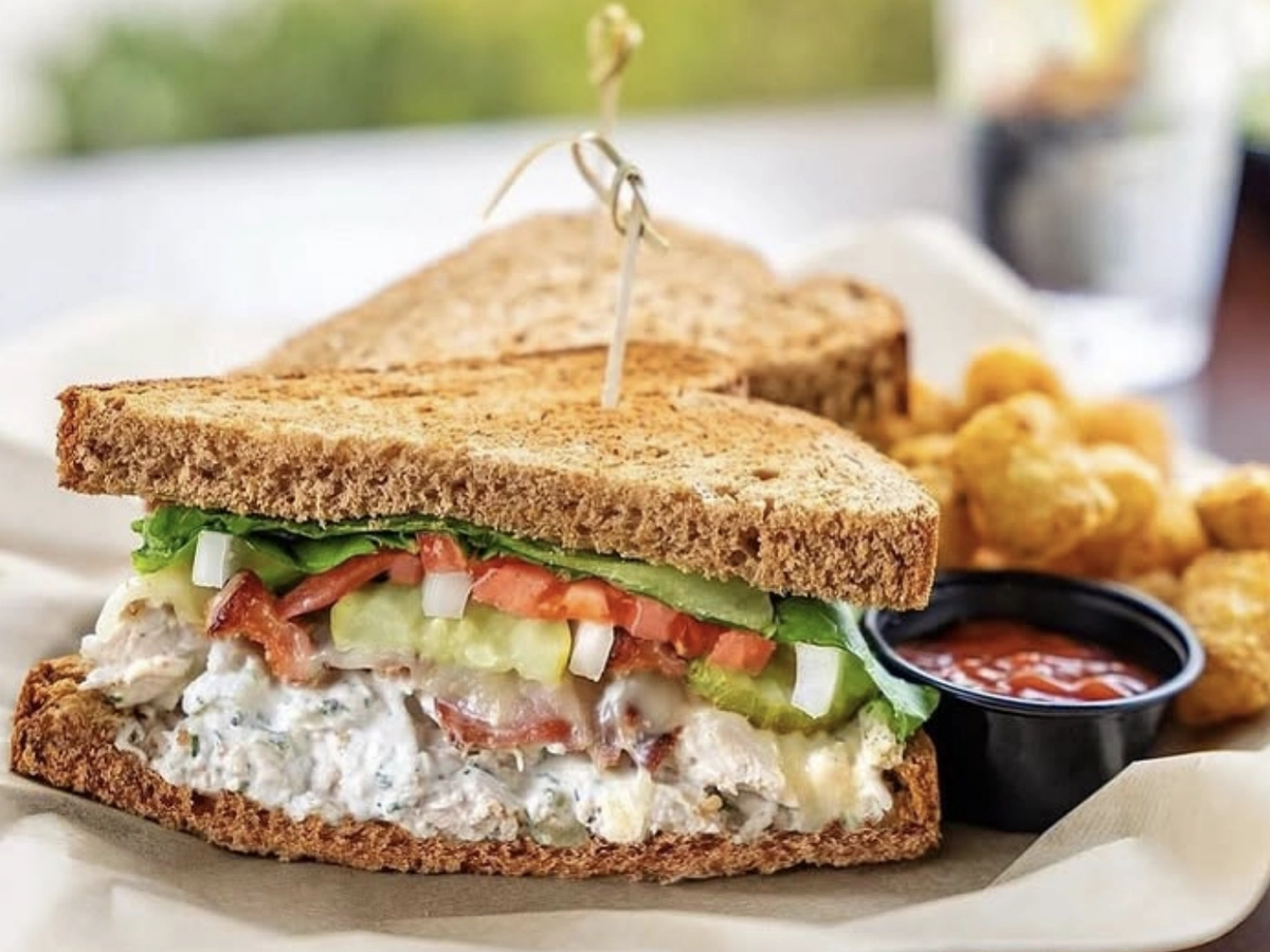 A hearty sandwich on toasted whole wheat bread filled with creamy chicken salad, lettuce, tomato, pickle, onion, and bacon, served with a side of crispy tater tots and ketchup.