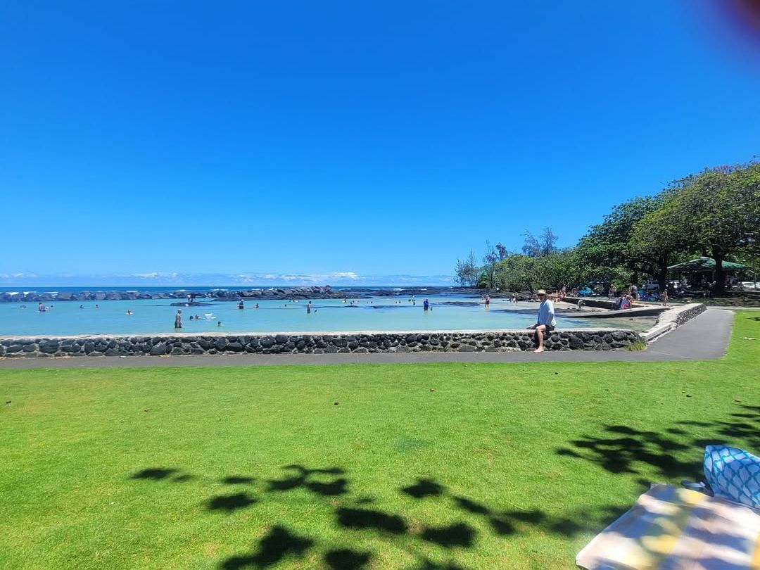 Families enjoy a sunny day at Onekahakaha Beach Park, a safe swimming spot with calm, shallow waters perfect for kids. The grassy picnic area and stone wall along the shoreline create a relaxing place to spend the afternoon.