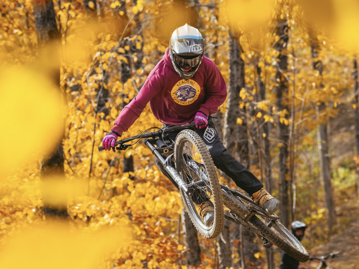 A mountain biker in a helmet and maroon sweatshirt catches air on a dirt trail surrounded by vibrant autumn foliage.