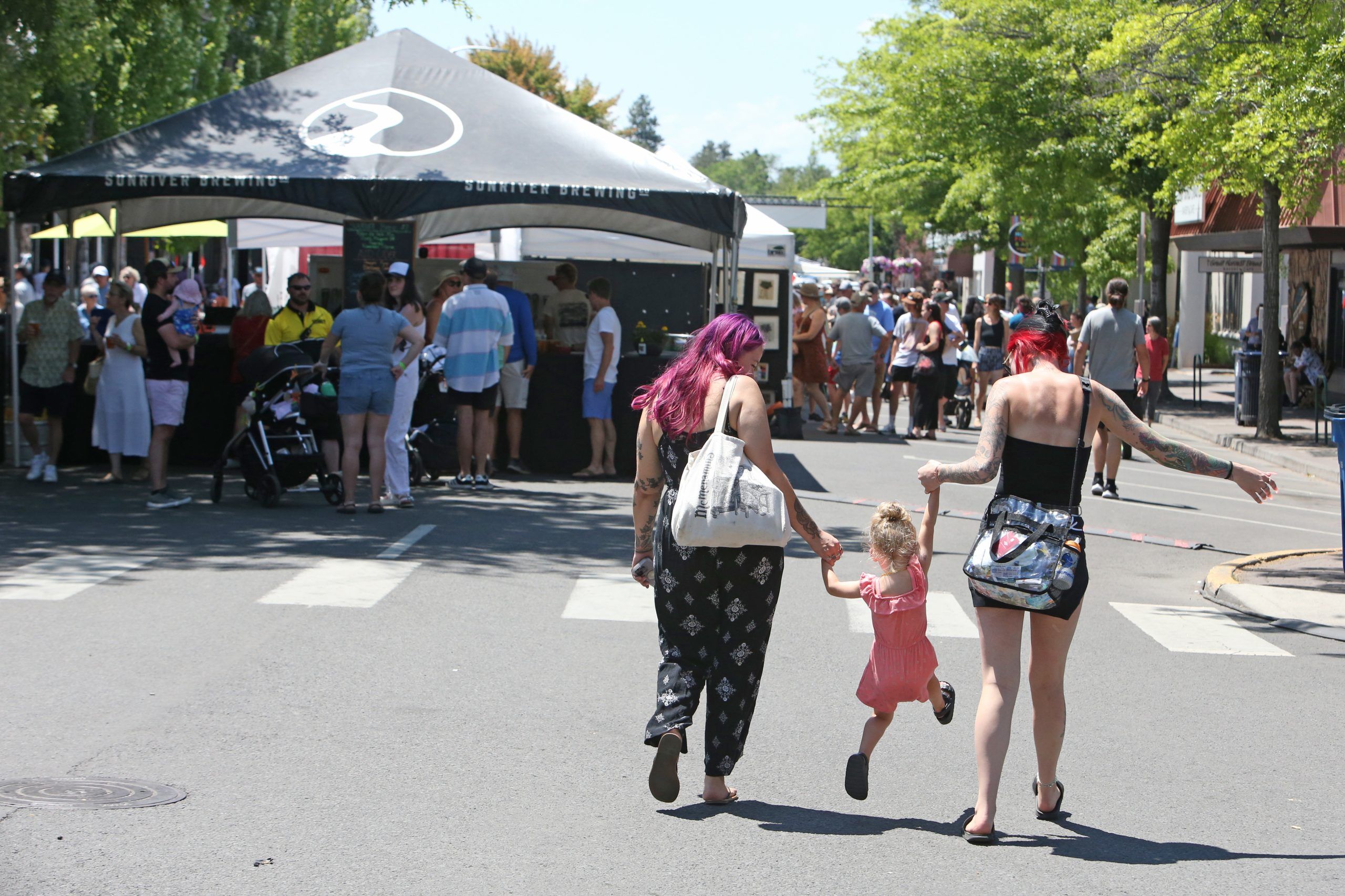 A cheerful summer crowd strolls through vendor booths on a tree-lined street as two adults swing a young child playfully between them. The warm weather, live music, and local food stands capture the joy of Bend’s Summer Festival.