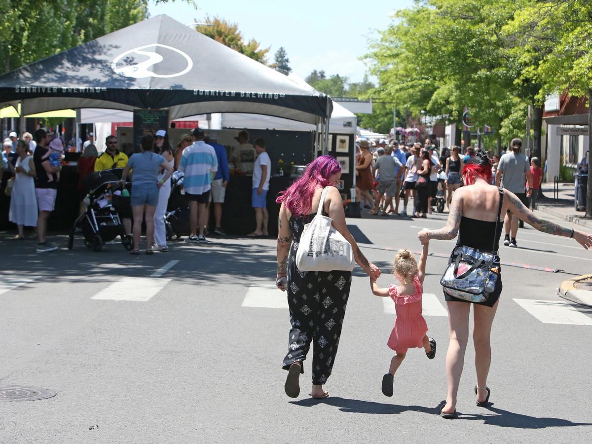 A cheerful summer crowd strolls through vendor booths on a tree-lined street as two adults swing a young child playfully between them. The warm weather, live music, and local food stands capture the joy of Bend’s Summer Festival.