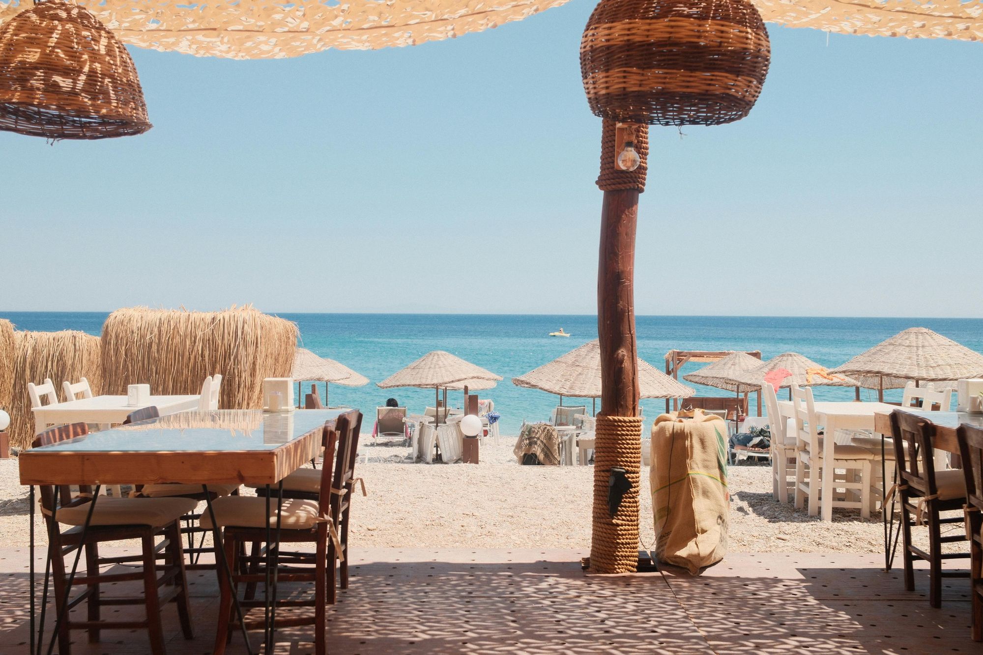 A breezy beachside restaurant with wooden tables, wicker lights, and views of straw umbrellas and turquoise waves. Perfect spot to enjoy a relaxed lunch just steps from the sand.