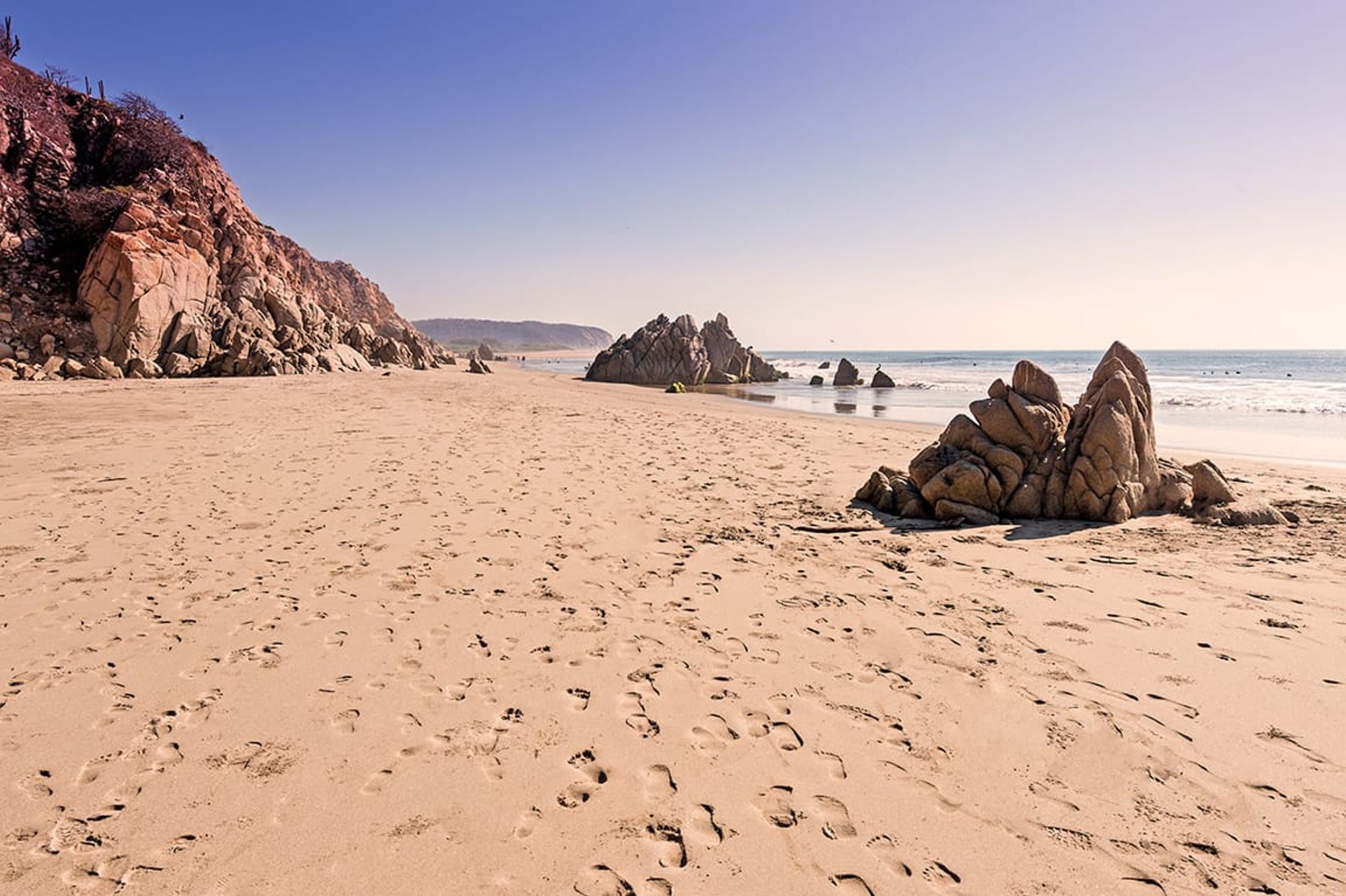 A quiet beach with soft sand and striking rock formations by the ocean, perfect for peaceful morning walks or sunset views. Footprints lead toward the waves, creating a calm and natural vibe.