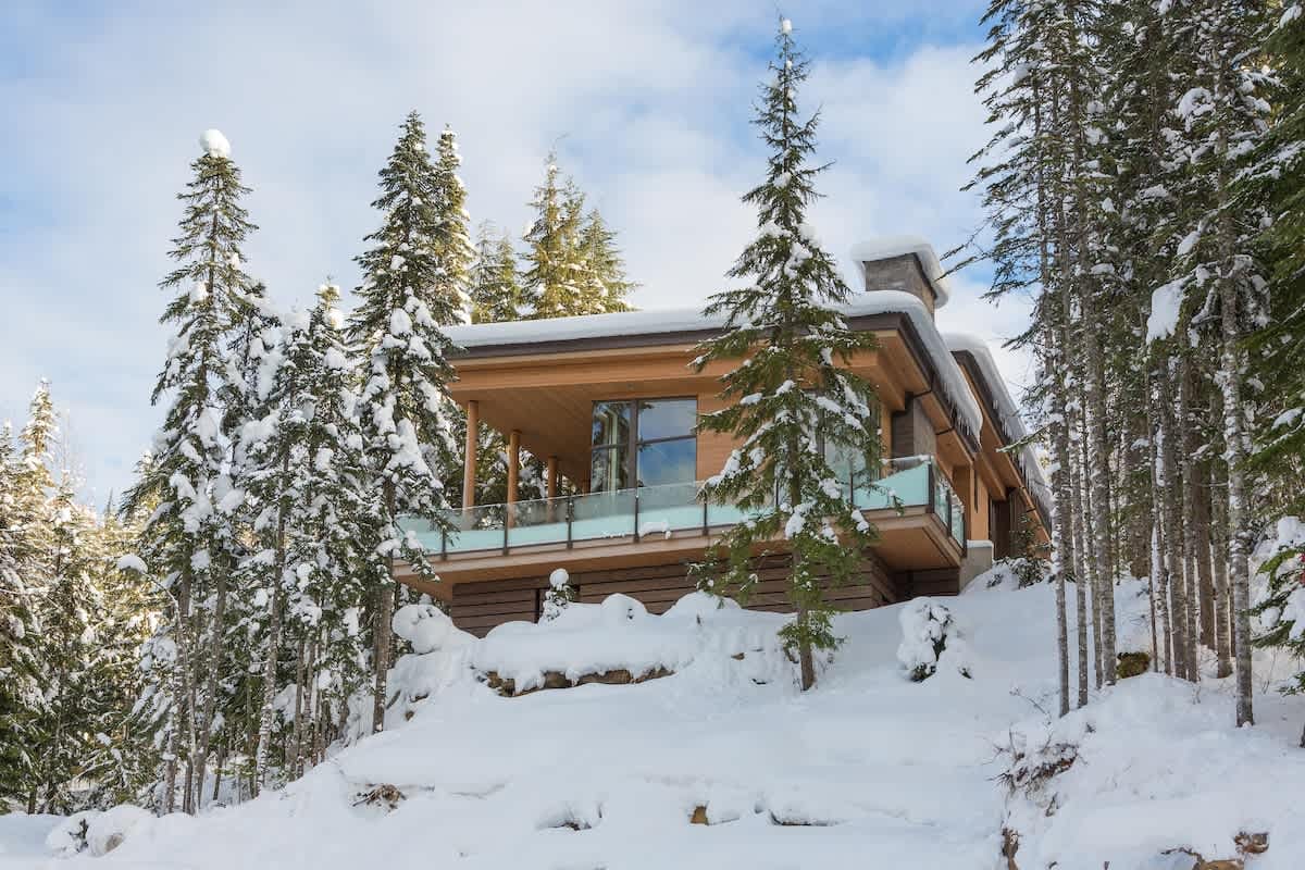 Modern mountain home with warm wood siding and glass balcony, perched on a snowy hillside surrounded by tall pine trees under a bright winter sky.