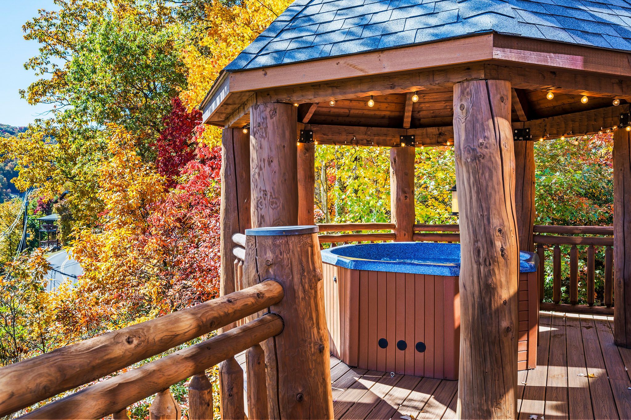 A cozy wooden gazebo with a hot tub inside, surrounded by vibrant autumn foliage in shades of red, orange, and gold, under a clear blue sky.