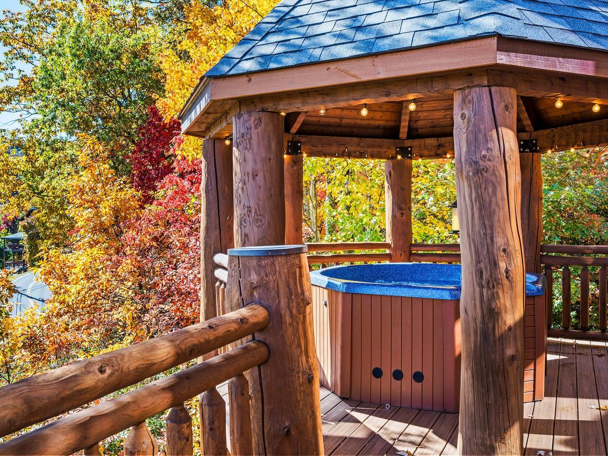 A cozy wooden gazebo with a hot tub inside, surrounded by vibrant autumn foliage in shades of red, orange, and gold, under a clear blue sky.
