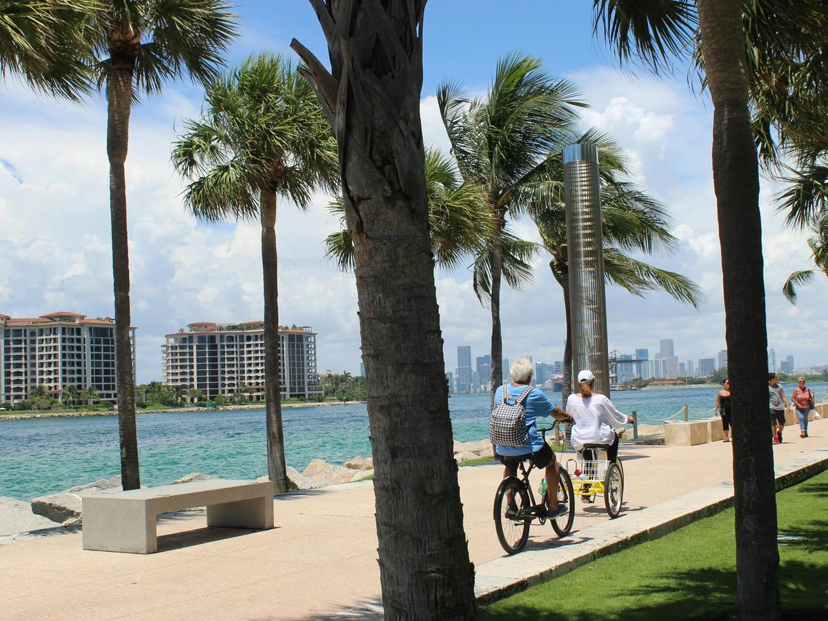 A couple enjoys a scenic bike ride along a palm-lined walkway beside turquoise waters. Modern beachfront condos rise in the distance, with the Miami skyline visible under a bright blue sky.