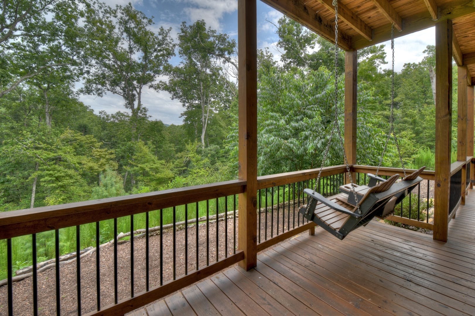 A covered wooden porch with a hanging bench swing overlooks a lush, green forest landscape.