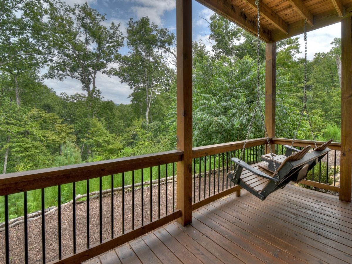 A covered wooden porch with a hanging bench swing overlooks a lush, green forest landscape.