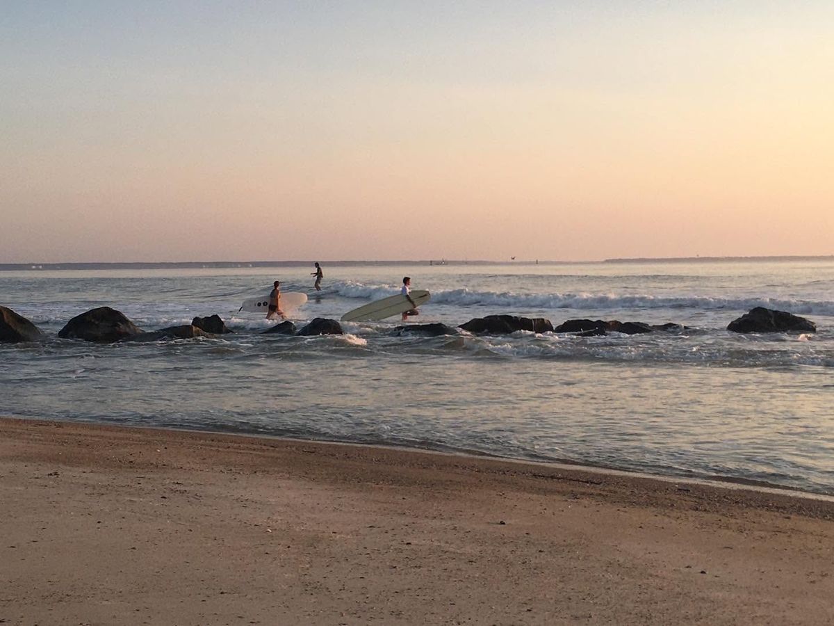 hree surfers carry their longboards across a rocky jetty in the shallow surf as the sky glows soft pink and gold at sunset. The calm ocean stretches toward a hazy horizon along Tybee Island's Mid Beach