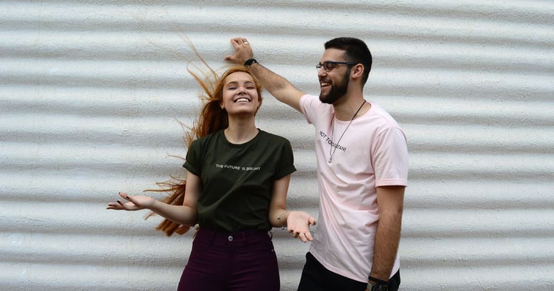 Two young people laughing and posing playfully against a white ribbed wall, like casual social media content.