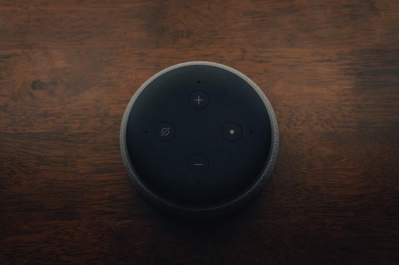 Top-down view of a dark Amazon Echo Dot smart speaker showing its four buttons on a wooden table.