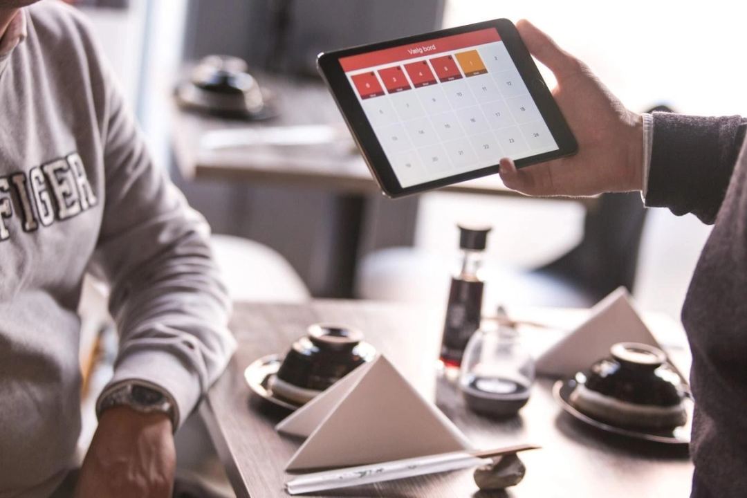 Person holding a tablet with a table-selection screen next to a seated diner at a table with soy sauce and folded napkins.