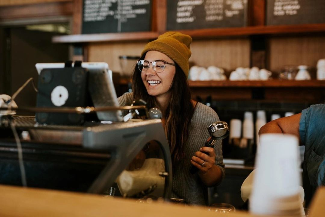 Café worker in glasses laughing behind the counter while holding an espresso portafilter, illustrating friendly service.
