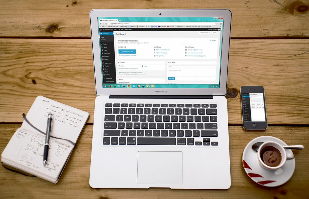 MacBook Air on a wooden desk showing the WordPress dashboard, beside a phone, a sketched notebook and a cup of coffee.